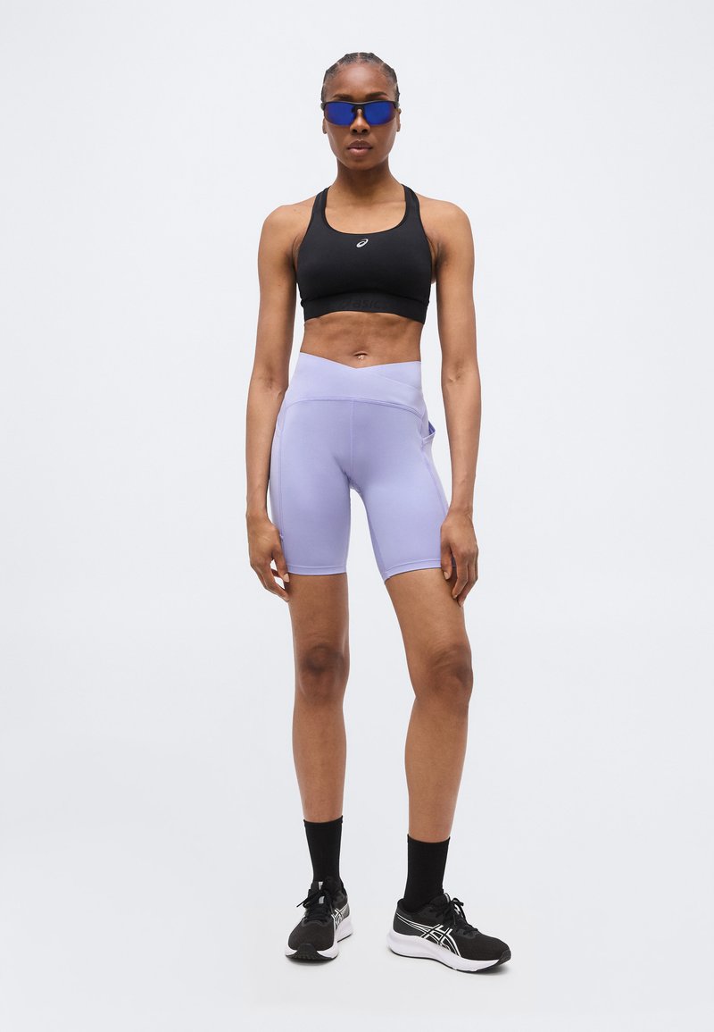 Atletische vrouw die een zwarte sportbeha, lichtpaarse fietsbroek, zwarte sokken, hardloopschoenen en reflecterende zonnebril draagt, staand tegen een effen achtergrond.