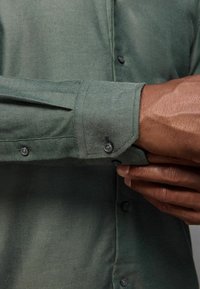 Eine dunkelhäutige Person, die einen Knopf am langen, olivgrünen Hemdärmel mit sichtbaren Knöpfen und strukturierter Stoffqualität zuknöpft.