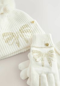 White knitted beanie and gloves adorned with gold sequined bow designs. Beanie features a fluffy pom-pom and a heart-shaped button detail.