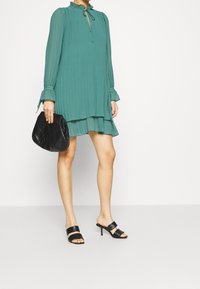 Turkusowa sukienka plisowana z falbankowym dekoltem, długimi rękawami i warstwowym dołem. Połączona z czarną pikowaną torebką i czarnymi klapkami na obcasie.