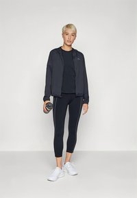 Melna rāvējslēdzēja jaka pār melnu krekliņu, kopā ar gludiem melniem legingiem ar baltām sānu svītrām un baltām sporta kurpēm. Tur rokā ūdens pudeli.