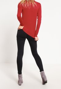 Un haut rouge à manches longues avec un ourlet arrière courbé, associé à un jean skinny noir et des bottines grises. Le tissu semble lisse et ajusté.