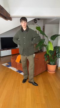 Noor mees, kes kannab rohelist jopet ja pruune pükse, seisab tubastes tingimustes oranži diivani ja suure taimepotiga puidust põranda peal.