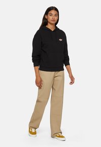 Felpa nera con cappuccio, tasca frontale e logo, abbinata a pantaloni beige a gamba larga e sneakers gialle con dettagli bianchi.