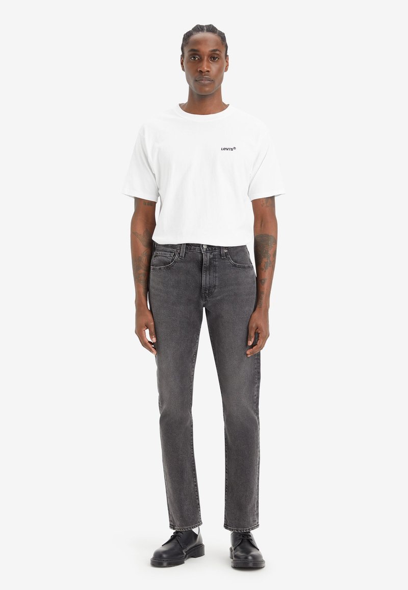 T-shirt blanc à manches courtes avec logo noir Levi's associé à un jean droit gris et des chaussures noires à lacets. Design minimaliste dans l'ensemble.