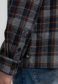 Flanellen shirt met een marineblauwe, grijze en oranje ruitpatroon, voorzien van zwarte knopen en een label aan de onderzijde aan de linkerkant.