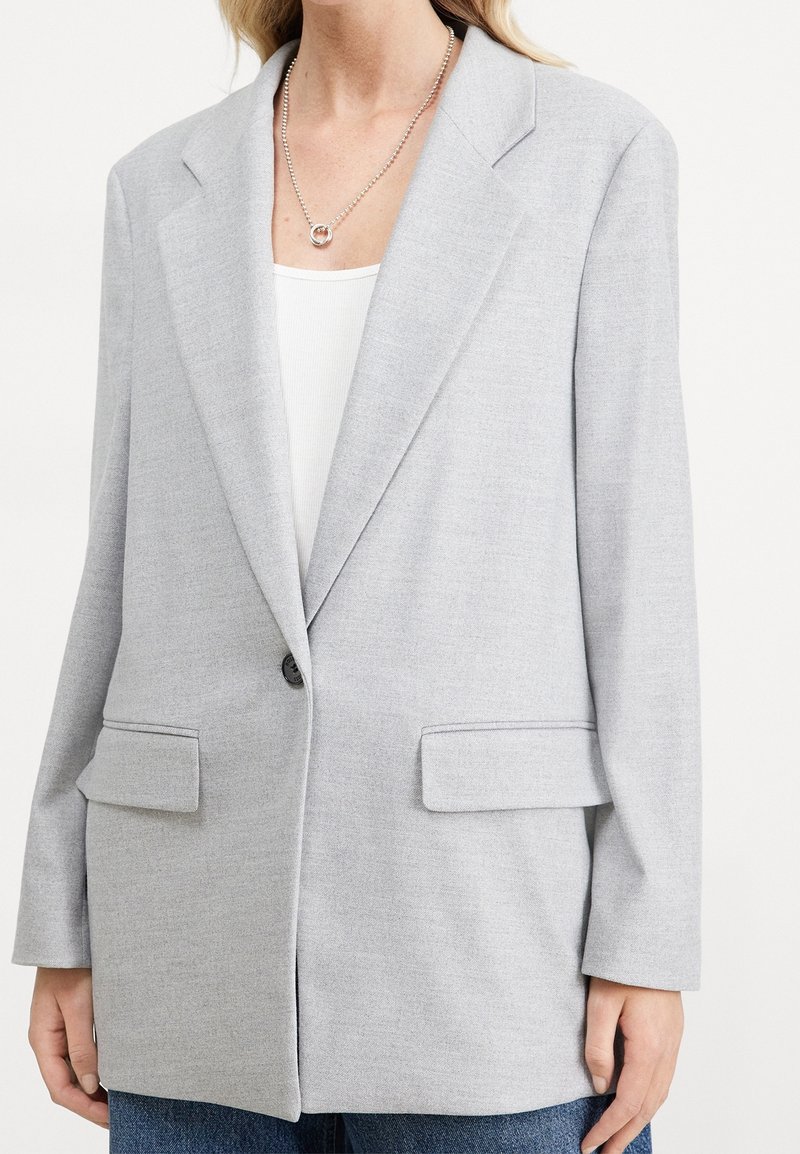 Blazer gris clair à coupe structurée, col à revers crantés, fermeture à un bouton, deux poches avant et tissu texturé subtil.