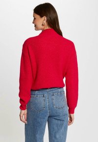 Pull en tricot rouge avec un col haut et une texture côtelée, associé à un jean bleu taille haute, vu de dos.