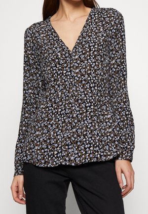 Kobieta ubrana w czarną bluzkę z długim rękawem ozdobioną drobnym niebiesko-białym kwiatowym wzorem oraz czarne spodnie, stojąca z rozluźnionymi rękami.