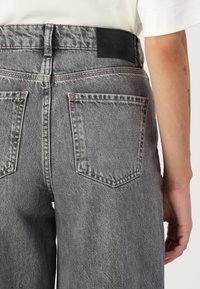 Harmaat farkut korkean vyötärön kanssa, suuret takataskut ja teksturoitu pinta. Vyötäröllä musta nahkainen brändipatch.