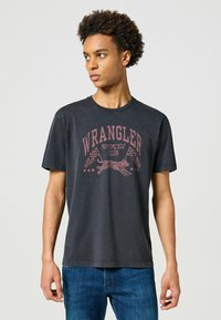 Camiseta de algodón negra con un gráfico rojo desvaído que incluye el texto "CLUB DE MOTOCICLETAS WRANGLER", un tigre y banderas a cuadros. Mangas cortas.