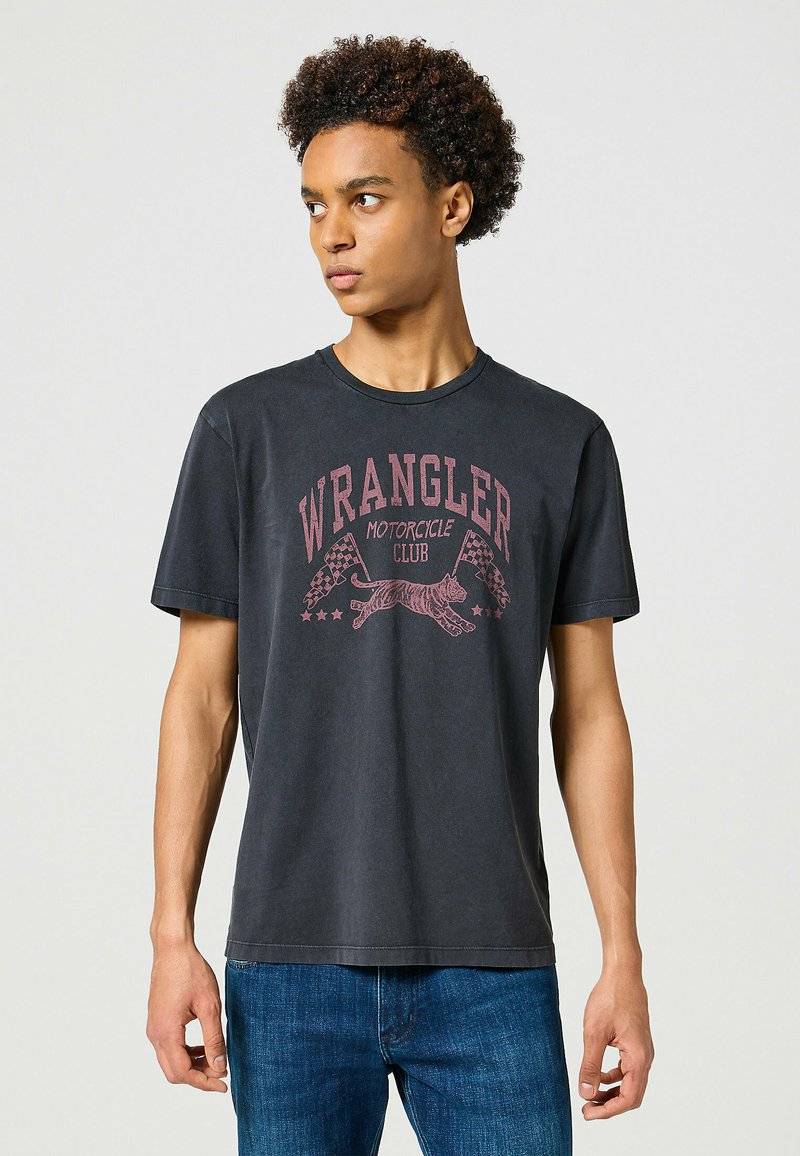 Camiseta de algodón negra con un gráfico rojo desvaído que incluye el texto "CLUB DE MOTOCICLETAS WRANGLER", un tigre y banderas a cuadros. Mangas cortas.