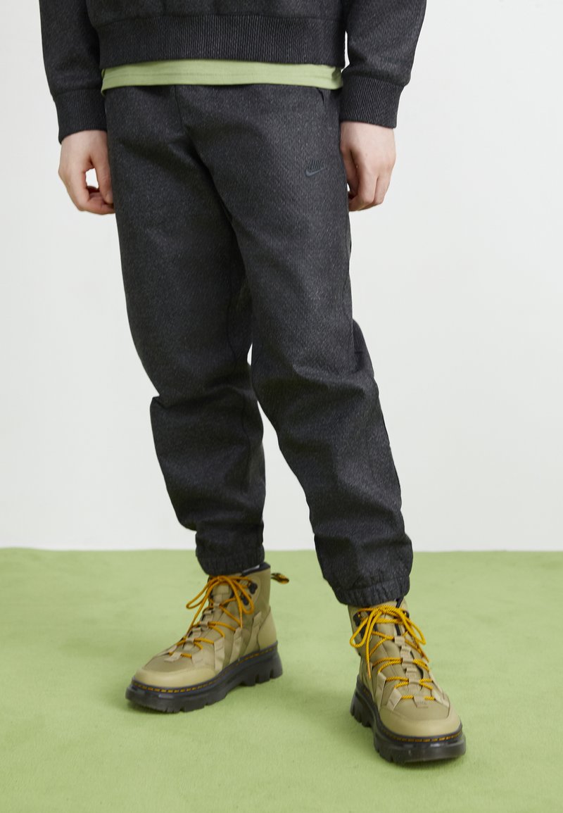 Pantalons de sport gris foncé avec des poignets élastiques, associés à des bottes beiges avec des lacets orange et une finition texturée. Fond vert.