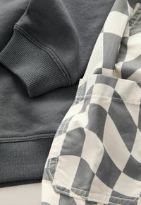 Zbliżenie na ciemnoszary ściągacz z prążkiem bluzy, nachodzący na szaro-białą materiał w kratkę, z widocznymi detalami ściegów.