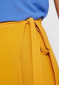 Gonna avvolgente gialla con una finitura testurizzata, dotata di una vita con nodo. La parte superiore è in tessuto blu. Dettaglio di cucitura visibile sulla gonna.