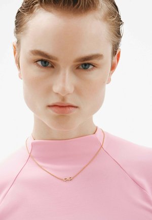 Collarín de oro con símbolo de infinito sobre una delicada cadena, llevando puesto sobre una blusa rosa suave de cuello alto. El fondo es blanco liso, lo que realza el enfoque en la joyería.