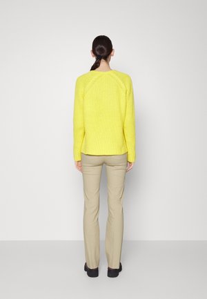 Kvinde stående med ryggen mod, iført en skriggul strikket sweater, beige bukser, sorte sko og med håret samlet i en hestehale.