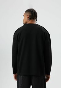 Schwarzes Langarmshirt aus strukturiertem Stoff, mit lockerem Schnitt und abgefallenen Schulternähten. Rückansicht zeigt ein schlichtes Design ohne Verzierungen.