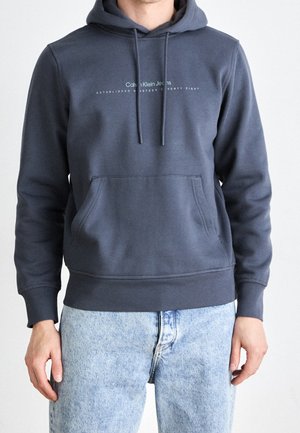 Homme portant un sweat à capuche Calvin Klein gris foncé et un jean bleu clair, debout, les bras détendus le long du corps, devant un fond uni.