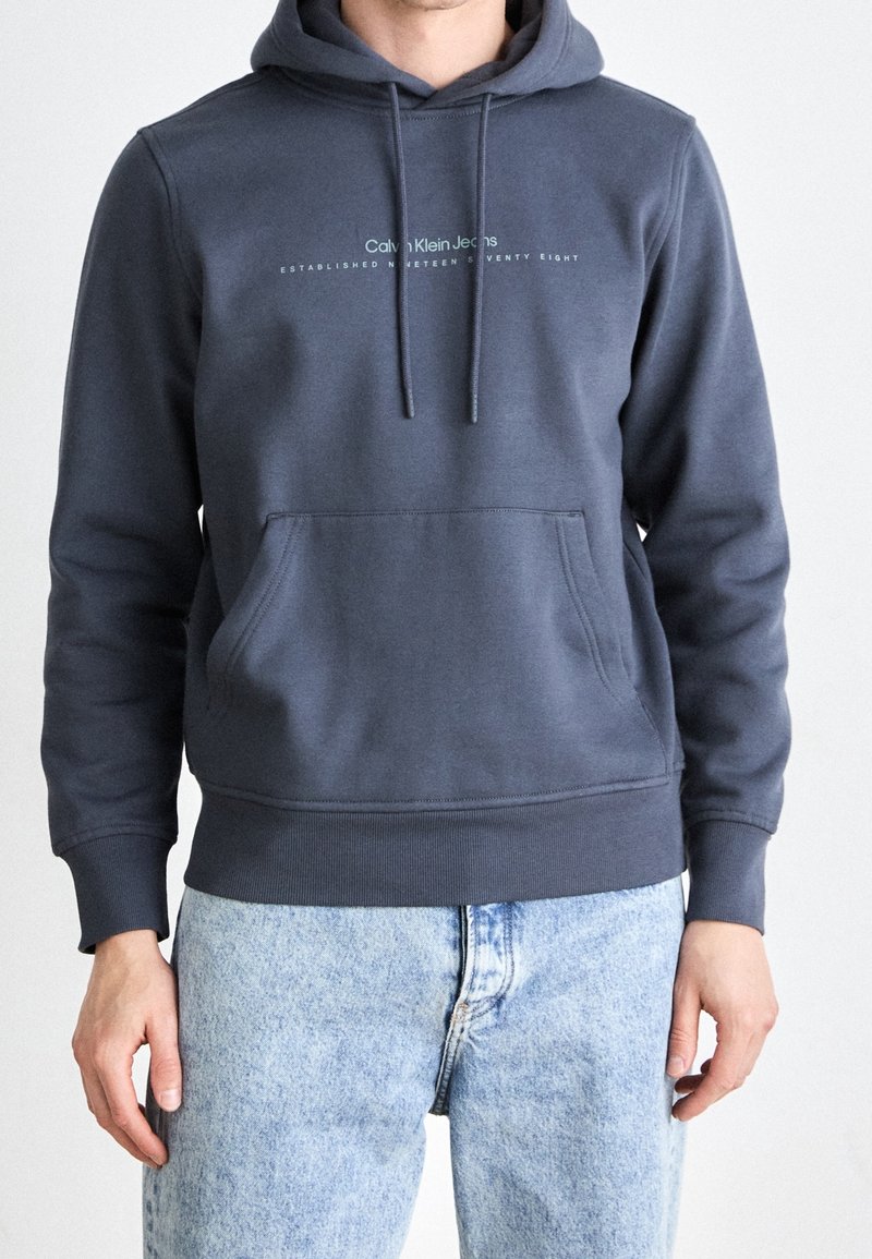 Homme portant un sweat à capuche Calvin Klein gris foncé et un jean bleu clair, debout, les bras détendus le long du corps, devant un fond uni.