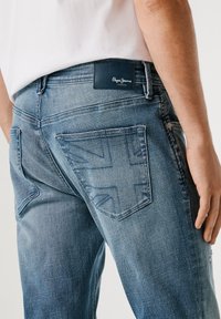 Vaqueros de mezclilla en azul claro, con un diseño en relieve de la bandera británica en el bolsillo trasero, y una etiqueta negra de "Pepe Jeans London" en la cinturilla.