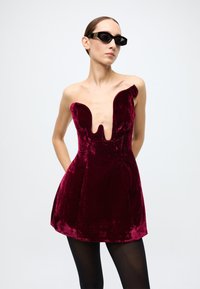 Burgundy velvet dress with a deep V-neckline, one shoulder design, flared skirt, and smooth texture. Worn with black sunglasses.