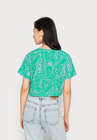 Top cropped verde com uma ilustração de inseto e planta brancos, mangas curtas, corte relaxado, combinado com jeans de ganga azul claro.