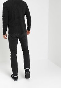 Svart långärmad skjorta med texturmönster och logotyp på ärmen, kombinerad med mörka denimjeans och svarta sneakers med vita accenter.