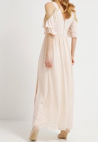 Largo vestido de un rosa pálido con diseño de hombros al aire, mangas con volantes y acento dorado en los hombros. Cuenta con un cierre de cremallera y aberturas laterales.