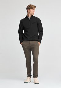 Svart zip-neck tröja i texturerat tyg, kombinerad med tanfärgade slim-fit byxor och gräddfärgade sneakers, accentuerad med gröna detaljer.