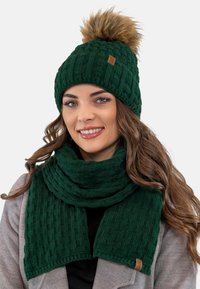 Berretto lavorato a maglia verde scuro con un pompon in pelliccia sintetica e una sciarpa coordinata. Entrambi presentano un motivo testurizzato e un piccolo logo decorativo.
