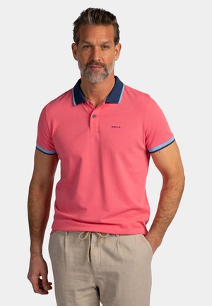 Man met grijze baard draagt een koraalroze poloshirt met blauwe kraag en manchetten, en beige broek met trekkoord, staand met één hand in de zak.
