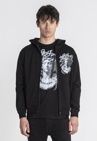 Crna zip-up dukserica s kapuljačom, s grafičkim printom klasične skulpture na prsima, nošena preko crne majice.