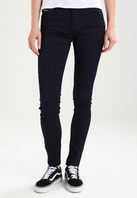 Jeans ajustados de color navy hechos de denim elástico, con cremallera y cinco bolsillos, estilizados con zapatillas negras y blancas.