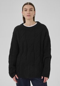 Frau in einem schwarzen, grob gestrickten Kabelpullover mit gerippten Bündchen und Saum, kombiniert mit dunkelblauen Jeans, steht vor einem schlichten, grauen Hintergrund.