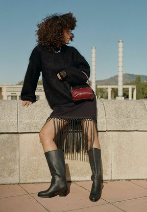 Suéter oversized negro, falda negra con flecos, botas negras hasta la rodilla y un pequeño bolso rojo. Fondo de concreto con cielo azul.