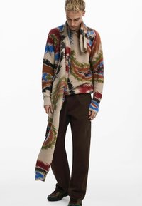 Cardigan a maglia a coste multicolore con motivi blu, verdi, rossi e beige, abbinato a pantaloni marroni e sneakers. Una lunga sciarpa scende.