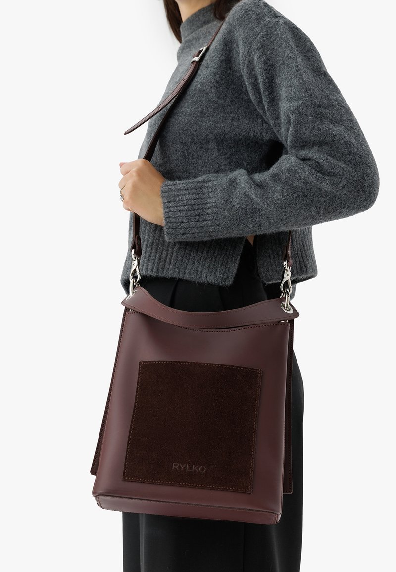 Brązowa skórzana torba na ramię z ciemnobrązową zamszową łatą, srebrnymi elementami metalowymi i regulowanym paskiem. Prostokątny kształt, gładka faktura.