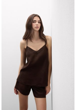 Jonge vrouw met lang bruin haar, gekleed in een donkerbruin mouwloos topje en bijpassende korte broek, staand tegen een effen witte achtergrond.