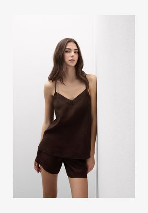 Jonge vrouw met lang bruin haar, gekleed in een donkerbruin mouwloos topje en bijpassende korte broek, staand tegen een effen witte achtergrond.