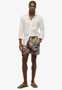 Camisa blanca de lino con botones, pantalones cortos navy con estampado floral, sandalias marrones y un reloj. Presenta hojas tropicales en colores vibrantes.