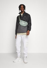 Sort vindjakke, lysegrønt crossover-axelremsväske, beige t-shirt, grå sweatpants med cargo-lommer, og hvide sneakers med lyse accenter.