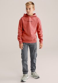 Sudadera con capucha roja y texto negro, combinada con pantalones cargo grises y zapatillas de deporte de un gris claro. Textura suave, ajuste relajado y cordón.