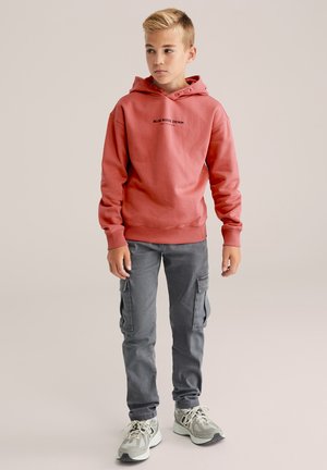 Rode hoodie met zwarte tekst, gecombineerd met grijze cargobroek en lichtgrijze sneakers. Gladde textuur, relaxte pasvorm en met trekkoord.