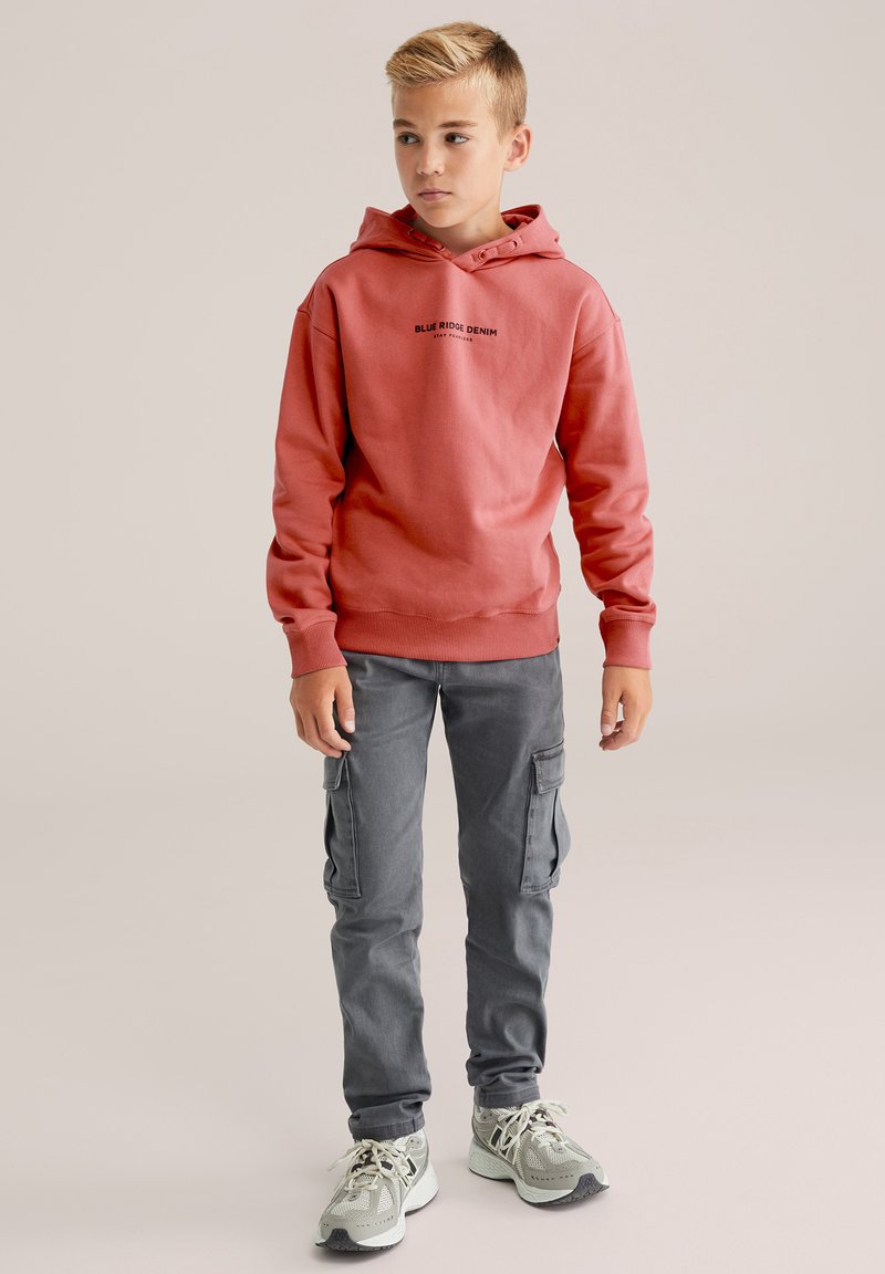 Sudadera con capucha roja y texto negro, combinada con pantalones cargo grises y zapatillas de deporte de un gris claro. Textura suave, ajuste relajado y cordón.