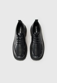 Un par de zapatos de cuero negro con cordones, suelas gruesas y costuras visibles, vistos desde arriba sobre un fondo claro.