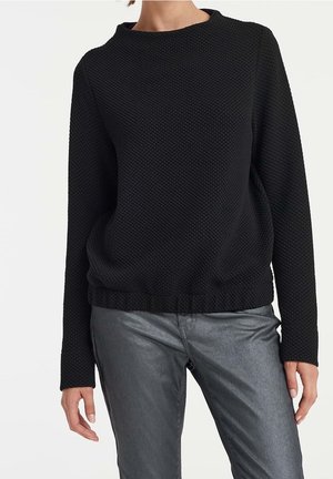 Kobieta ubrana w teksturowany czarny sweter z długim rękawem i metalicznie szare spodnie, stojąca na tle gładkiego białego tła.