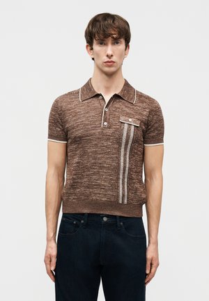 Άνδρας που φοράει καφέ πλεκτό πόλο με κοντά μανίκια, τσέπη στο στήθος και κάθετες λευκές ριγέ διακοσμήσεις, σε συνδυασμό με σκουρόχρωμα τζιν.