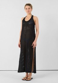 Robe maxi en tricot noir avec un design sans manches, un motif à mailles ouvertes, des fentes latérales et une coupe ample. Portée par-dessus un maillot de bain avec des tongs.