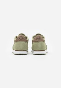Paire de baskets en daim vert avec semelles blanches vue de dos, avec des patches marron texturés au talon ornés de logos circulaires.